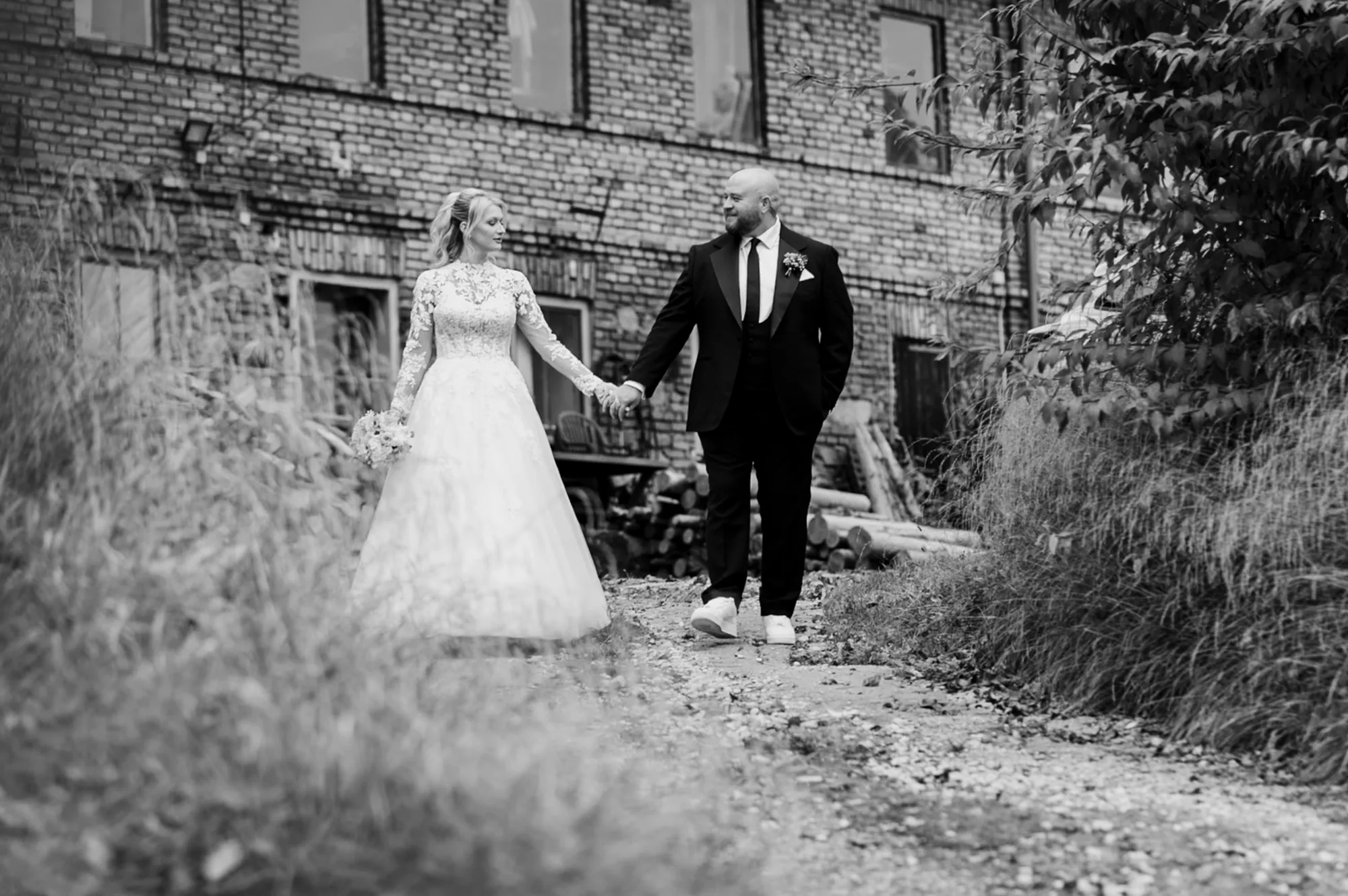 First-Look-Moment: Brautpaar geht Hand in Hand an einem alten Gutshof vorbei, Schwarz-Weiß